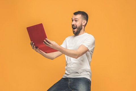 Young man with a beard looking at a red laptop with an excited expression on his face.