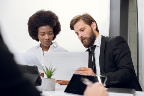 stock photo of creative young business people in formal wear, Caucasian man and African woman, sitting at the table at office brainstorming meeting and discussing their project, looking at papers together