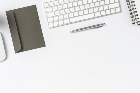 A plant, wireless mouse, grey envelope, keyboard, pen, and touchpad on a white desk.