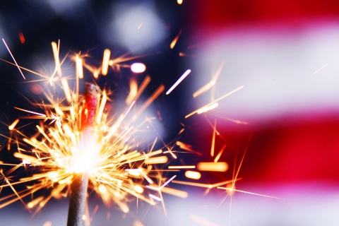 Photograph of a sparkler in action with an American flag in the background, representing fireworks on the fourth of July