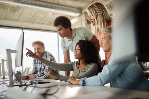 A group of five business people looking at a computer together at an agency, collaborating on the design of insurance agency websites and discussing the launch of a new website.