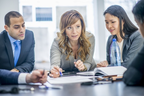 Stock photo of a group of professionals sitting around a conference table engaged in a discussion. They are reviewing documents representing collaboration between top insurance aggregators and independent agents within an insurance agency setting. The scene conveys teamwork and strategic planning in the insurance industry.